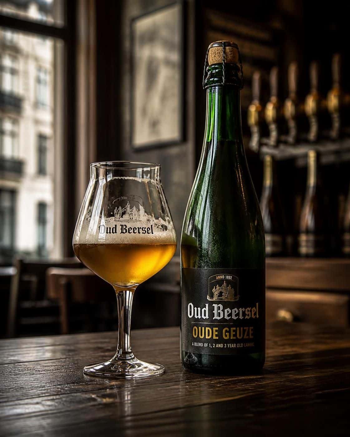 Bouteille verte d’Oud Beersel et verre de bière dorée sur une table près d’une fenêtre